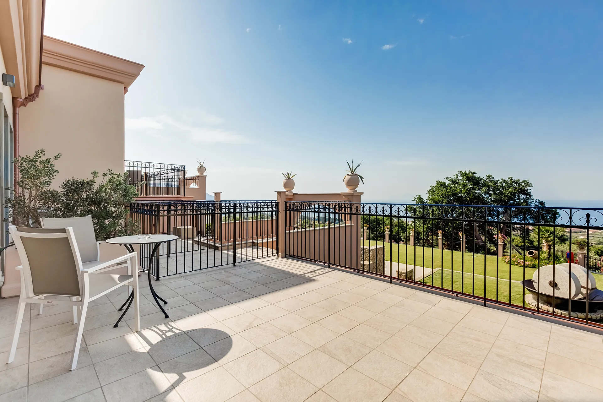 Terrazza dell’hotel con sedie e tavolino bianchi, parapetto in ferro e vista sul giardino e sul mare.