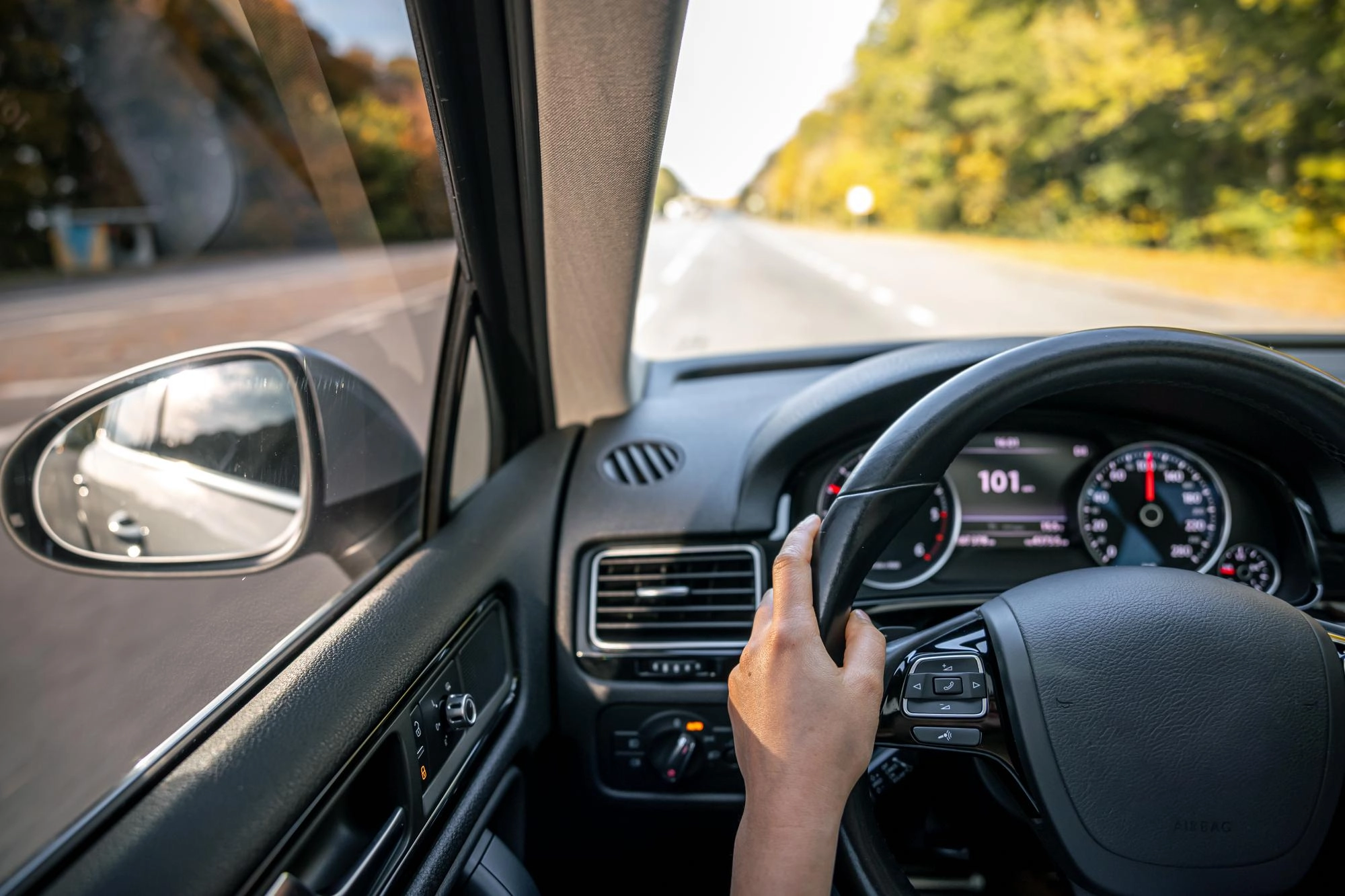 Vista dall’interno di un’auto in movimento su una strada, con una mano sul volante e paesaggio alberato sullo sfondo