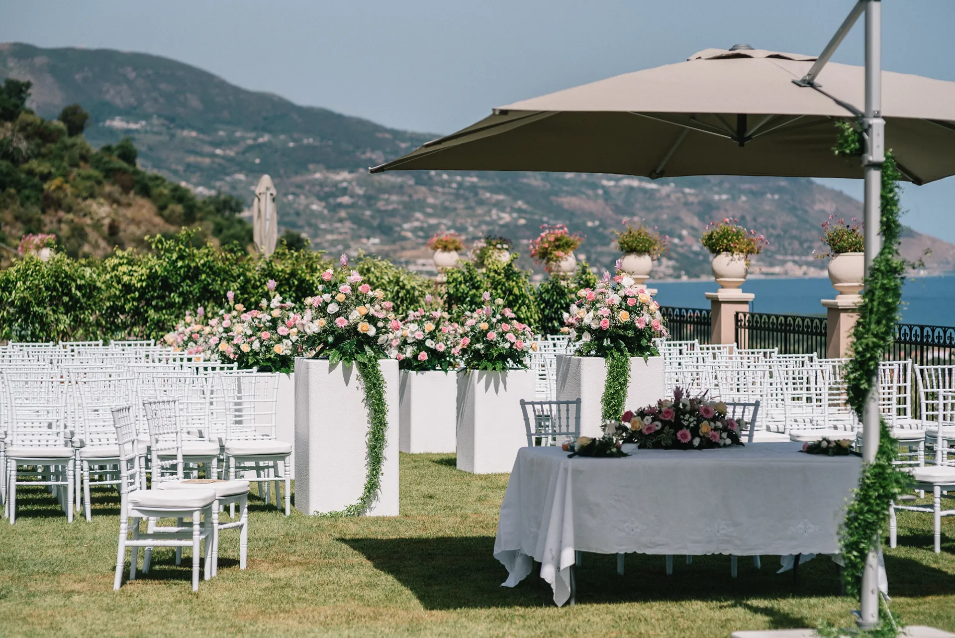 Giardino panoramico dell’Hotel Terre di Eolo a Patti allestito per una cerimonia all’aperto, con sedie bianche, composizioni floreali su piedistalli e vista sulle colline e sul mare.