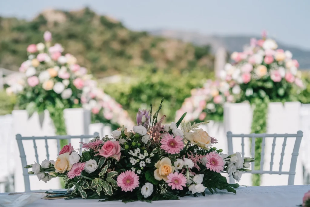 Centrotavola floreale con rose e gerbere rosa su tavolo per cerimonia all’aperto presso l’Hotel Terre di Eolo a Patti, con composizioni fiorite e colline sullo sfondo