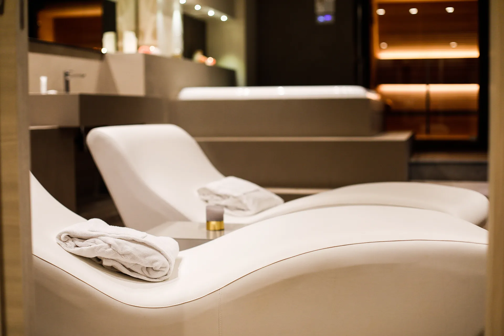 Zona relax dell’Hotel Terre di Eolo a Patti con lettini ergonomici bianchi, asciugamano piegato e vista sulla sauna illuminata, in un ambiente wellness moderno e confortevole.